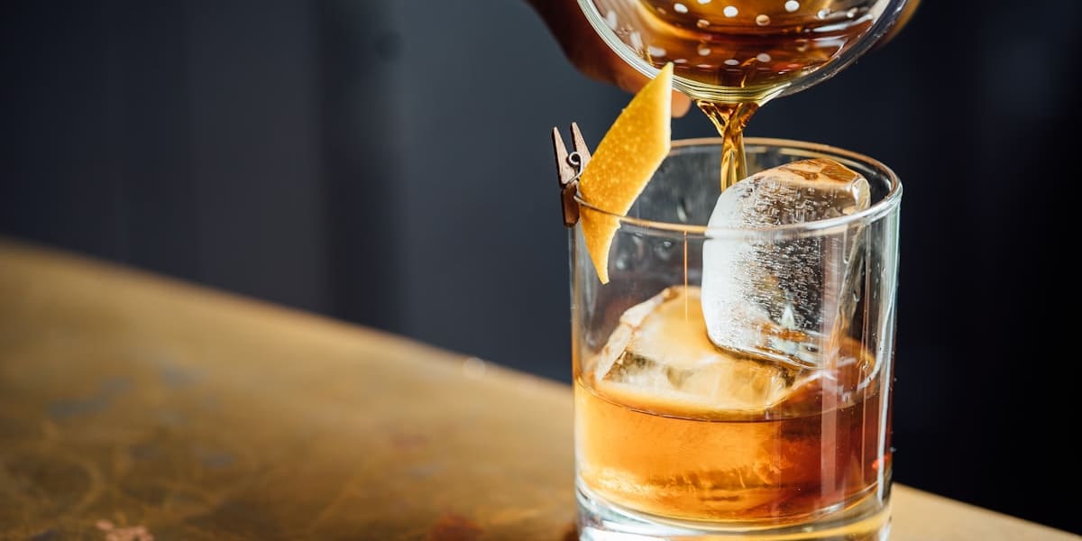 An amber cocktail being strained into a rocks glass with a large ice cube and an orange peel garnish on a worn bar counter.