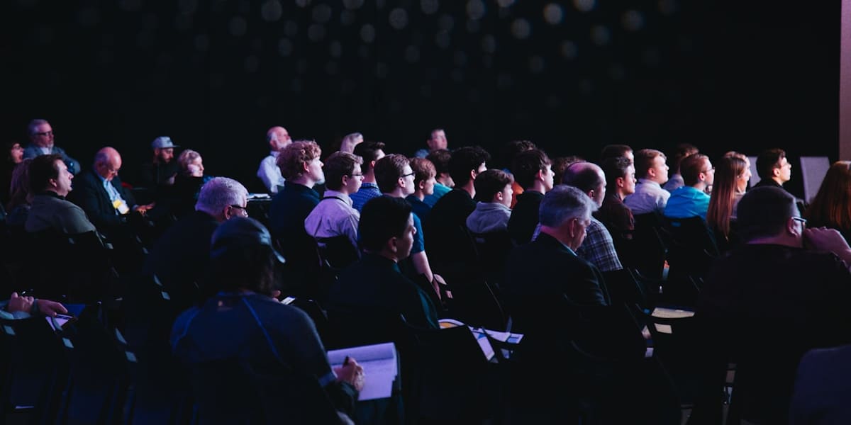 Rows of seated attendees watching a presentation in a dark conference hall with patterned wall lighting