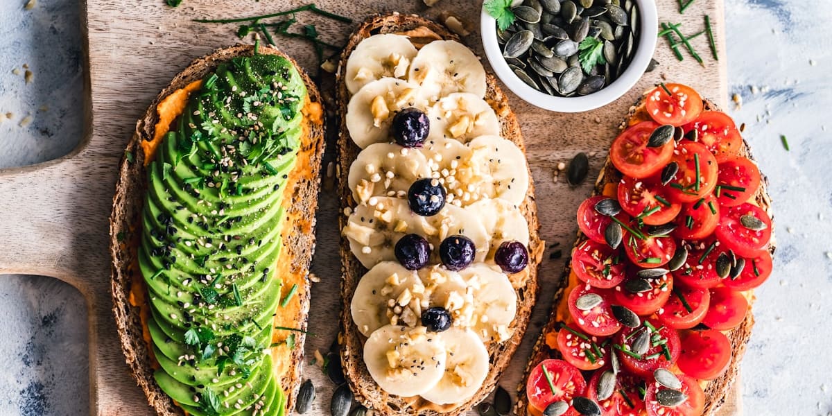 Three slices of toast topped with avocado, sliced banana and blueberries, and cherry tomatoes, with a bowl of pumpkin seeds