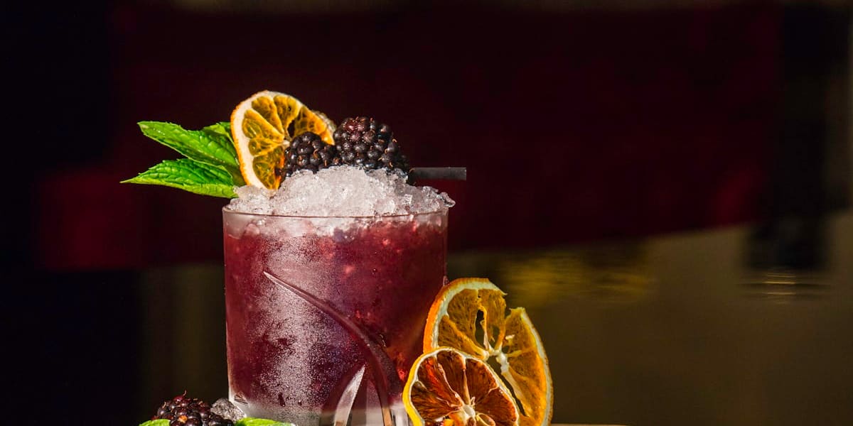 A dark blackberry cocktail topped with crushed ice, fresh blackberries, mint and a dried citrus wheel on a wooden board.
