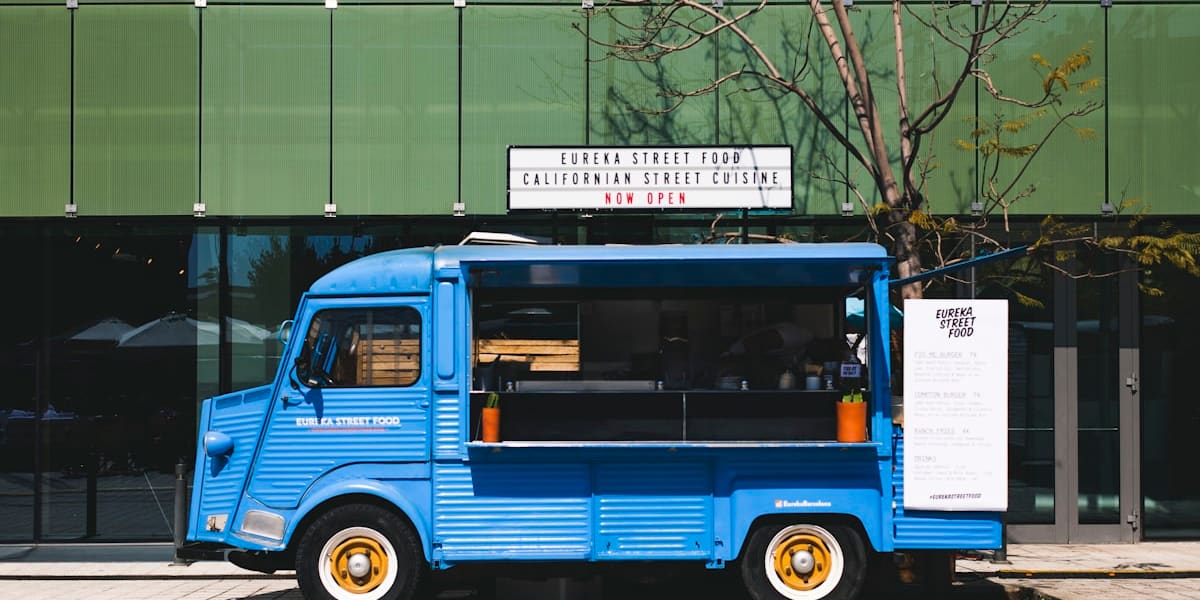 A blue vintage food truck with a hand-painted menu sign parked on a city sidewalk in front of a green glass building.