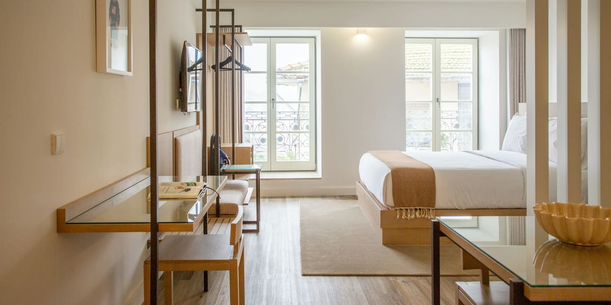 Bright, clean hotel room with a made bed, desk, light wood floor and tall windows