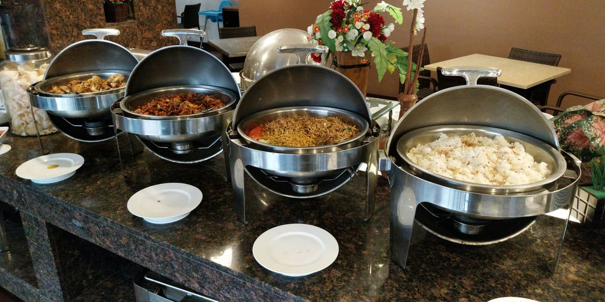 A row of open stainless steel chafing dishes holding hot food on a buffet counter