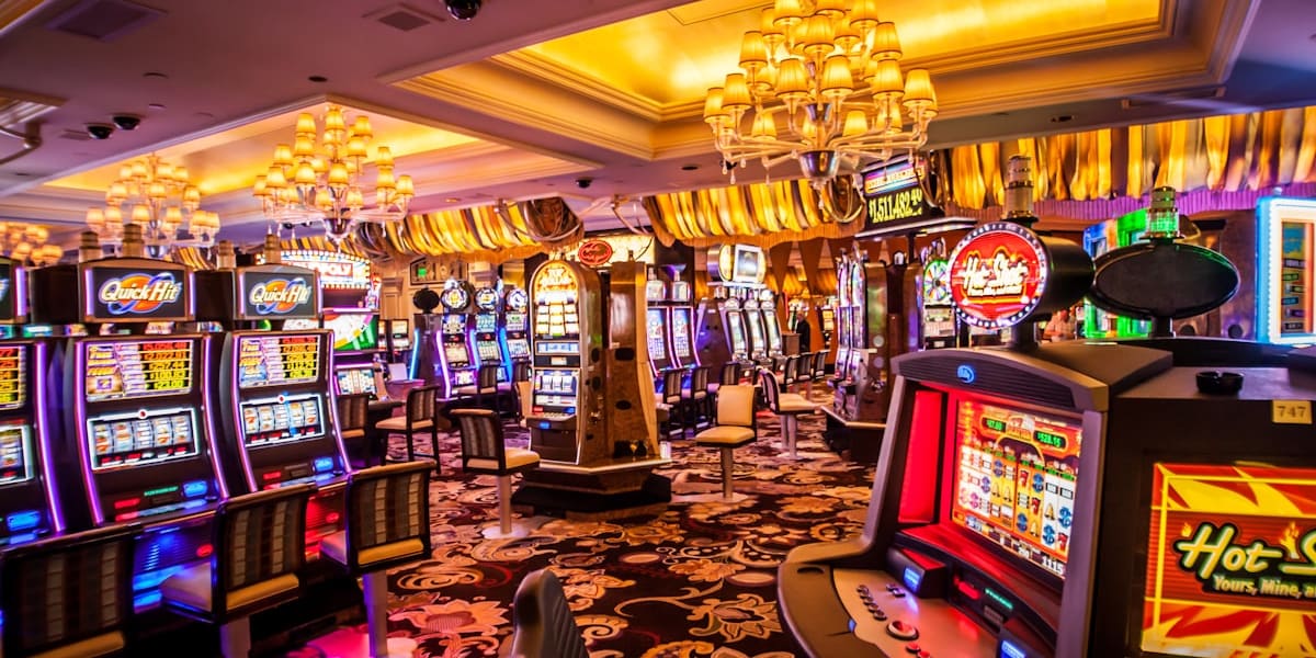 Casino floor lined with lit slot machines under ornate gold chandeliers and patterned carpet