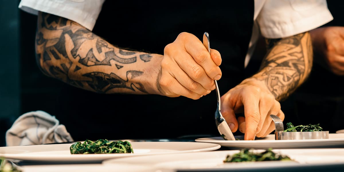 A chef with tattooed forearms plating cooked greens onto white dishes with a spoon