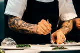 A chef with tattooed forearms plating cooked greens onto white dishes with a spoon