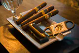 Three cigars resting on a white ashtray beside a cigar cutter and lighter in warm light