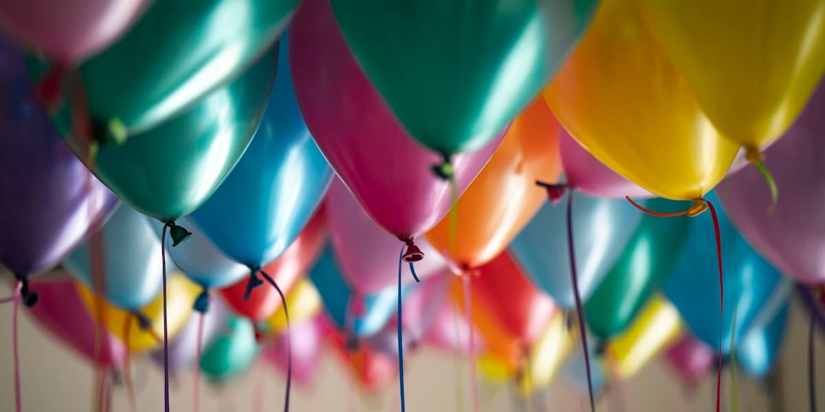 Cluster of colorful metallic party balloons with trailing ribbons against a plain ceiling