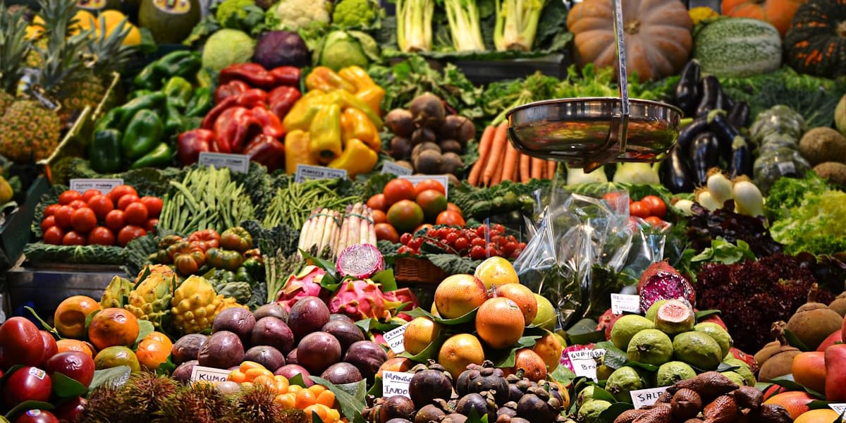 A crowded market stall piled high with colorful fruits and vegetables, including peppers, tomatoes and tropical fruit, with handwritten price signs.
