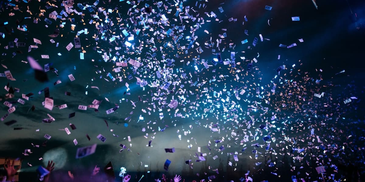 A concert crowd with arms raised under a shower of falling confetti, lit by blue and white stage lighting.