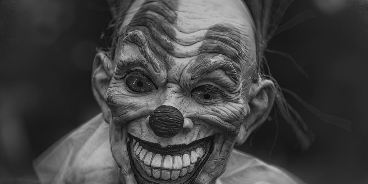 A black-and-white close-up of an unsettling wrinkled clown mask with a wide toothy grin and dark hollow eyes.