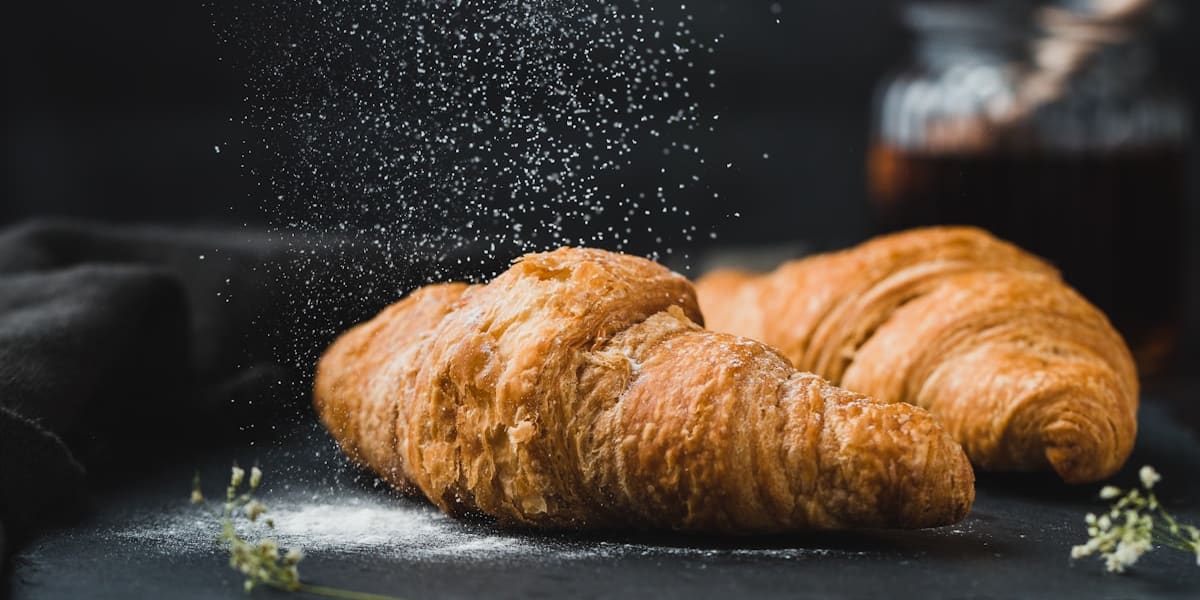 Two fresh croissants on a dark slate board being dusted with powdered sugar, with a honey jar behind