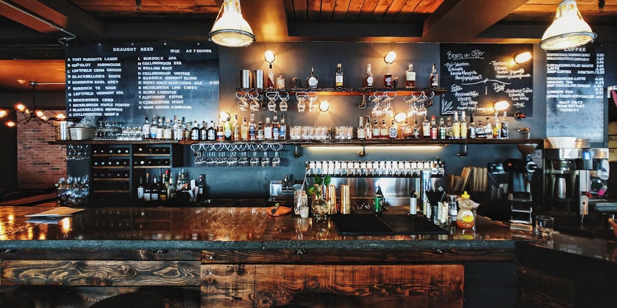 Dimly lit bar counter with shelves of liquor bottles, hanging glassware and wooden stools
