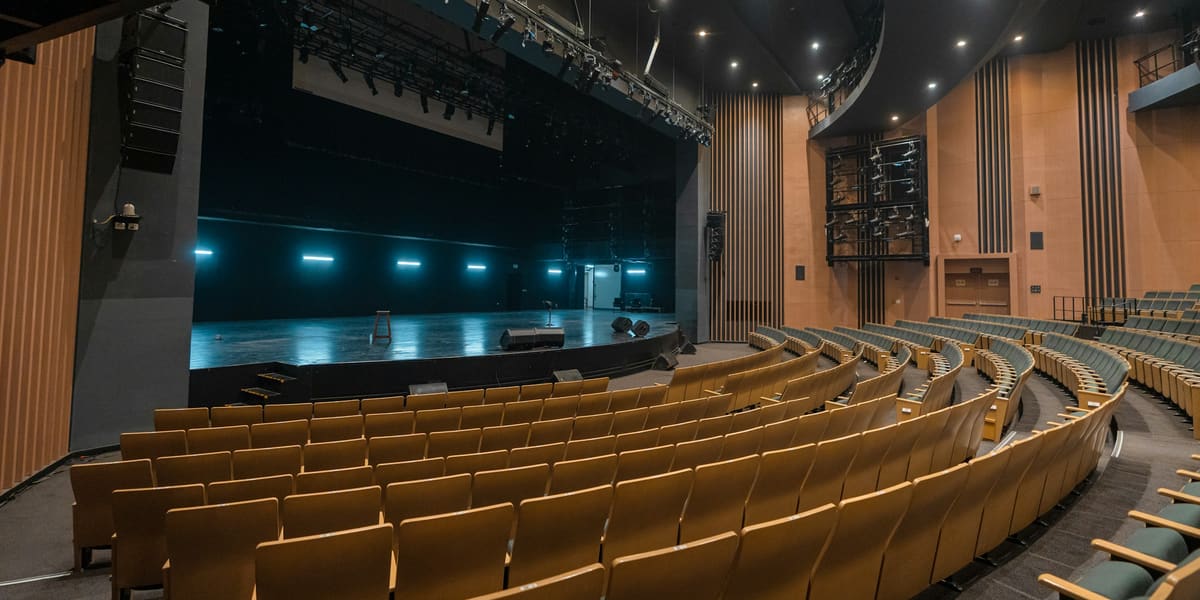 Curved rows of empty wooden theater seats facing a stage lit with blue lights