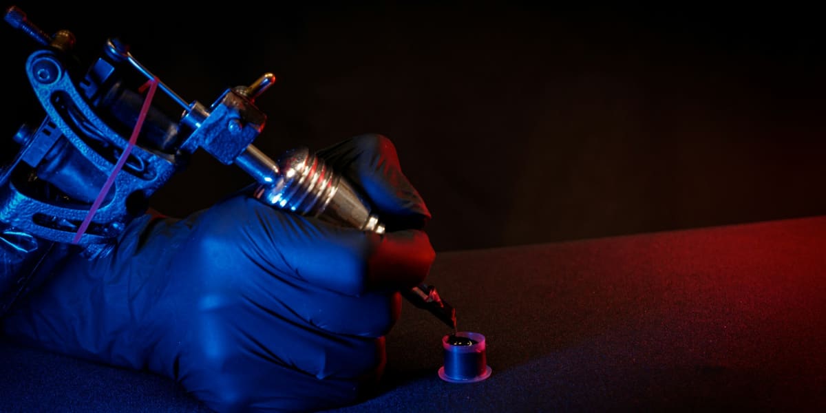 Gloved hand holding a coil tattoo machine over an ink cup under blue and red light