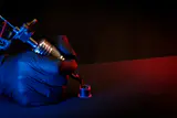 Gloved hand holding a coil tattoo machine over an ink cup under blue and red light