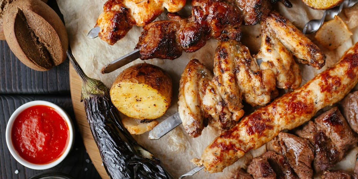 Overhead spread of grilled meat skewers, chicken wings, roasted vegetables and dipping sauces