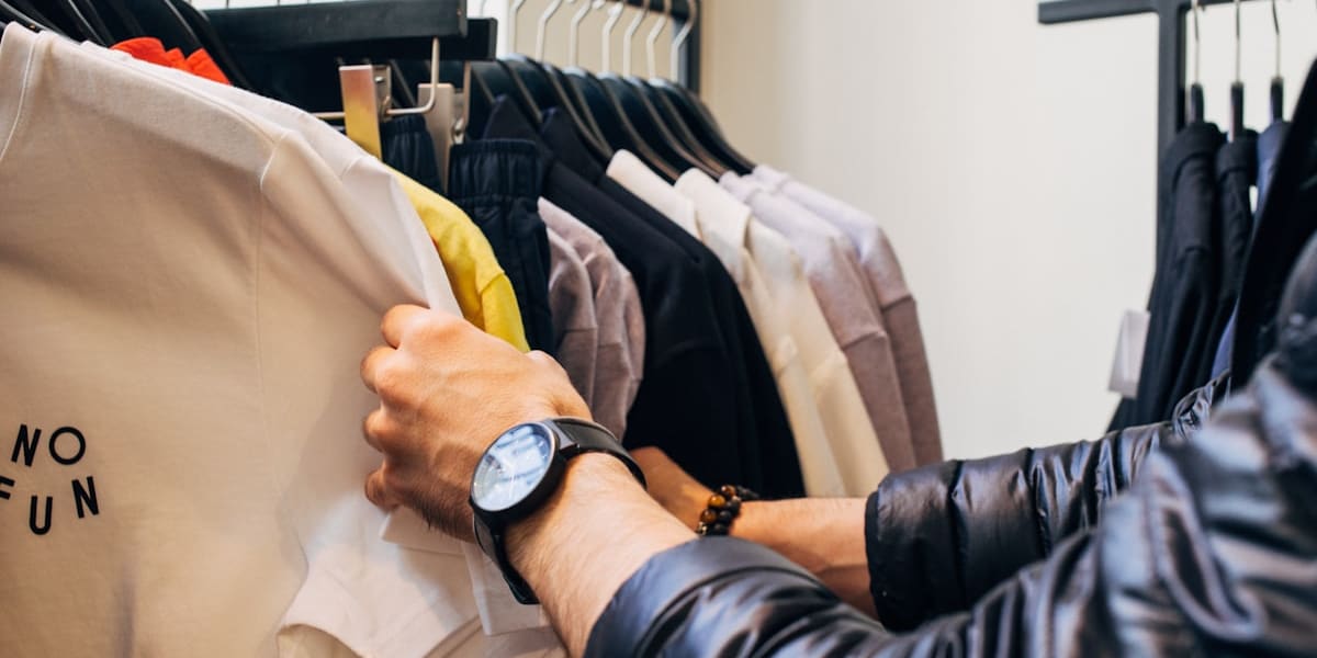 A hand sorting through a rack of folded t-shirts and hoodies on hangers inside a clothing store.