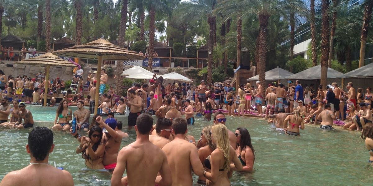 Crowd standing in shallow water at a daytime pool party under palm trees and thatched umbrellas