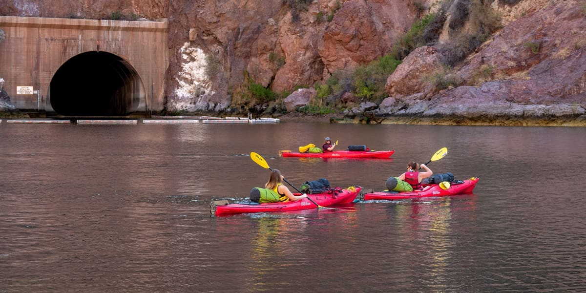 Three people paddling red kayaks on calm water beneath reddish canyon walls and a tunnel portal