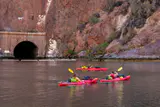 Three people paddling red kayaks on calm water beneath reddish canyon walls and a tunnel portal