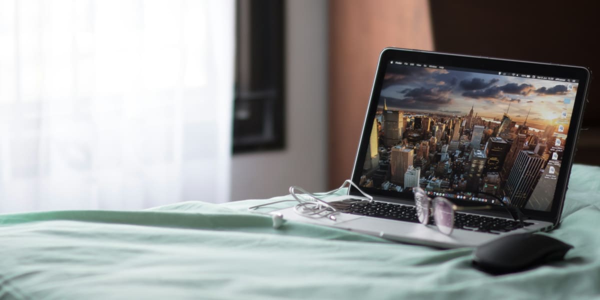 Open laptop with glasses and a mouse resting on a made bed beside a sunlit window