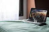 Open laptop with glasses and a mouse resting on a made bed beside a sunlit window