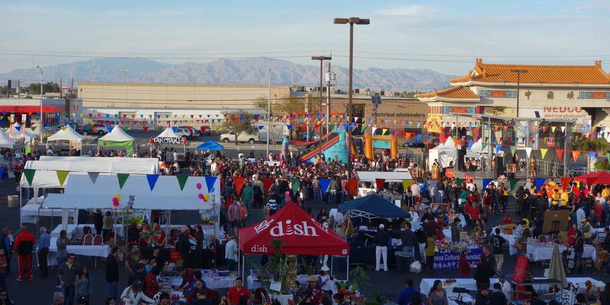 Crowds among food stalls and tents in the Chinatown Plaza parking lot, with pagoda-style roofs and mountains beyond