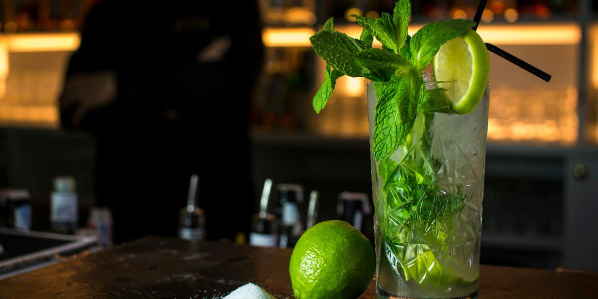 Mojito with mint and lime in a cut-glass tumbler on a dark bar counter, bartender blurred behind