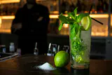 Mojito with mint and lime in a cut-glass tumbler on a dark bar counter, bartender blurred behind