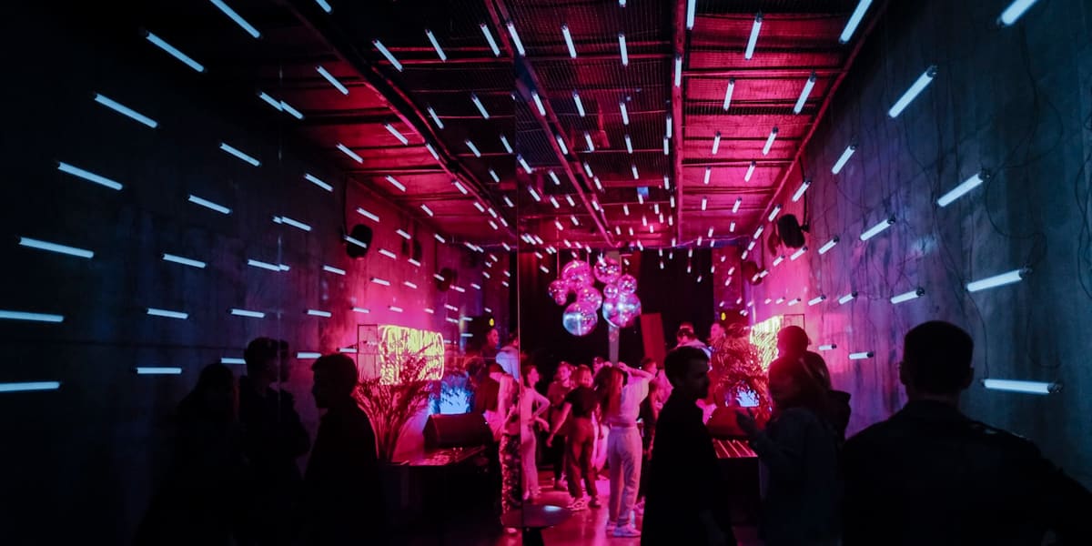 A nightclub corridor lined with glowing red and white neon tube lights on the ceiling and walls, with a crowd dancing below a disco ball.