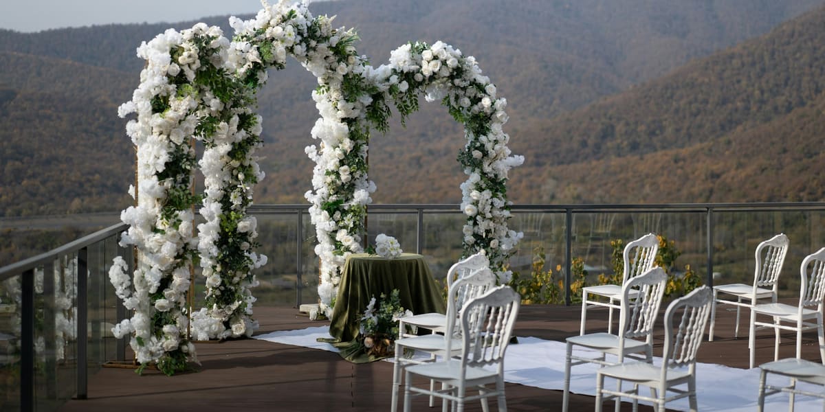 Two white floral arches and rows of white chairs set for an outdoor wedding on a terrace facing mountains