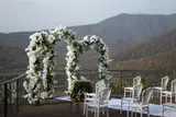 Two white floral arches and rows of white chairs set for an outdoor wedding on a terrace facing mountains
