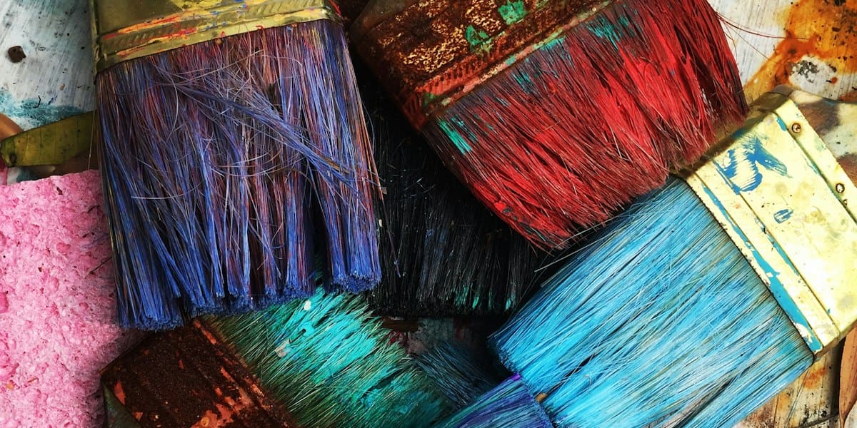 Several paintbrushes coated in bright blue, red, and purple paint resting on a paint-splattered surface