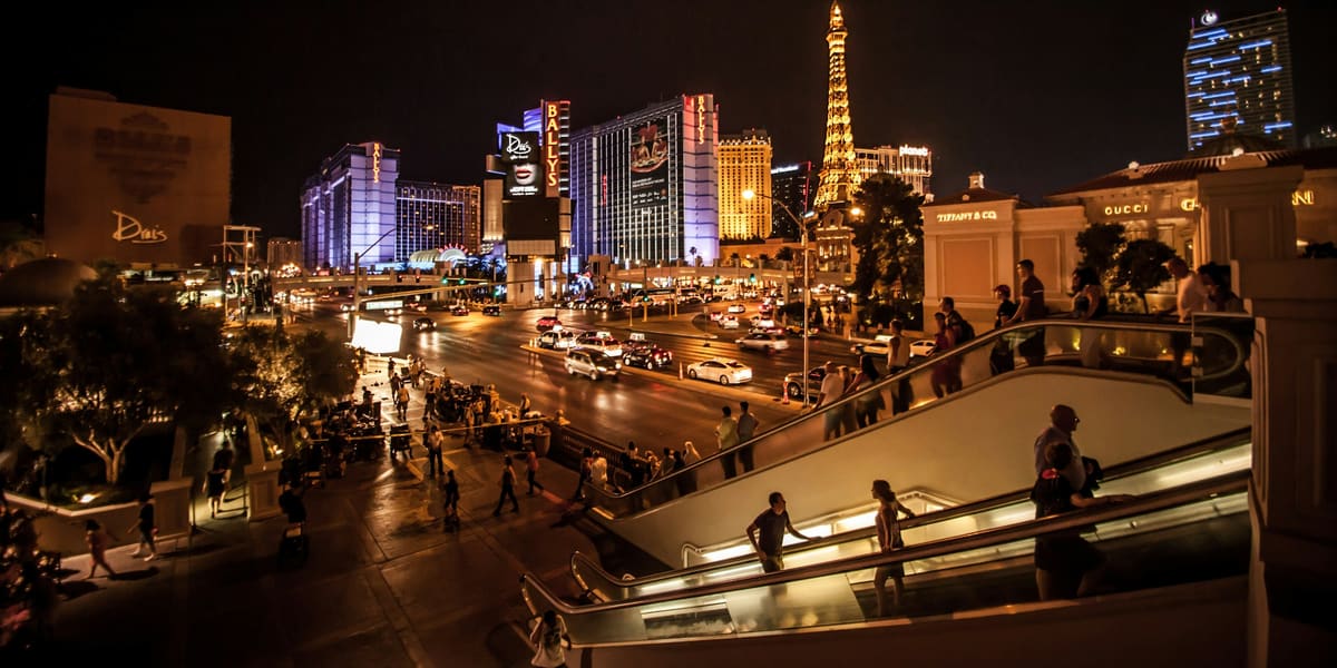 Pedestrians on escalators and sidewalks beside Las Vegas Boulevard at night, with the Paris tower lit beyond