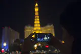 Hands holding up a smartphone to photograph a lit Eiffel Tower replica on the Las Vegas Strip at night