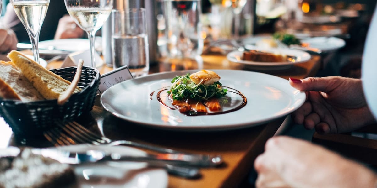 A small plated dish with greens and sauce on a restaurant table, surrounded by wine glasses, a water bottle and a bread basket.