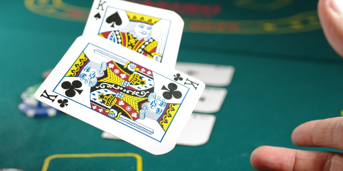 A hand holding two king playing cards above a green felt poker table with stacked chips and more cards in the background.