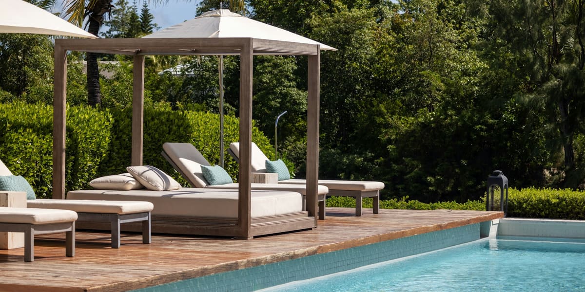 Wooden poolside cabana with a cushioned daybed and loungers beside a turquoise pool, palms and trees behind