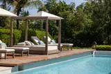 Wooden poolside cabana with a cushioned daybed and loungers beside a turquoise pool, palms and trees behind