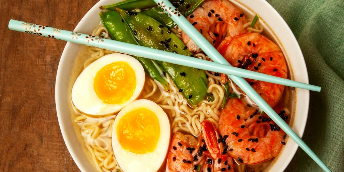 An overhead view of a ramen bowl with noodles, shrimp, soft-boiled egg halves and snow peas, topped with black sesame seeds.