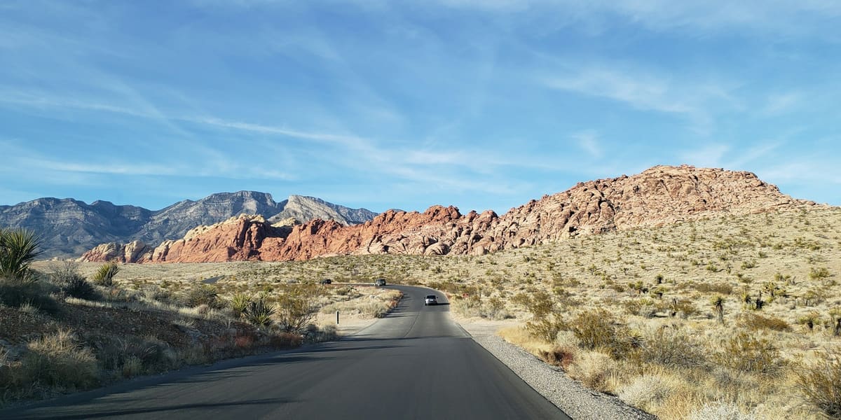Paved road curving through desert scrub toward red and tan sandstone hills at Red Rock Canyon