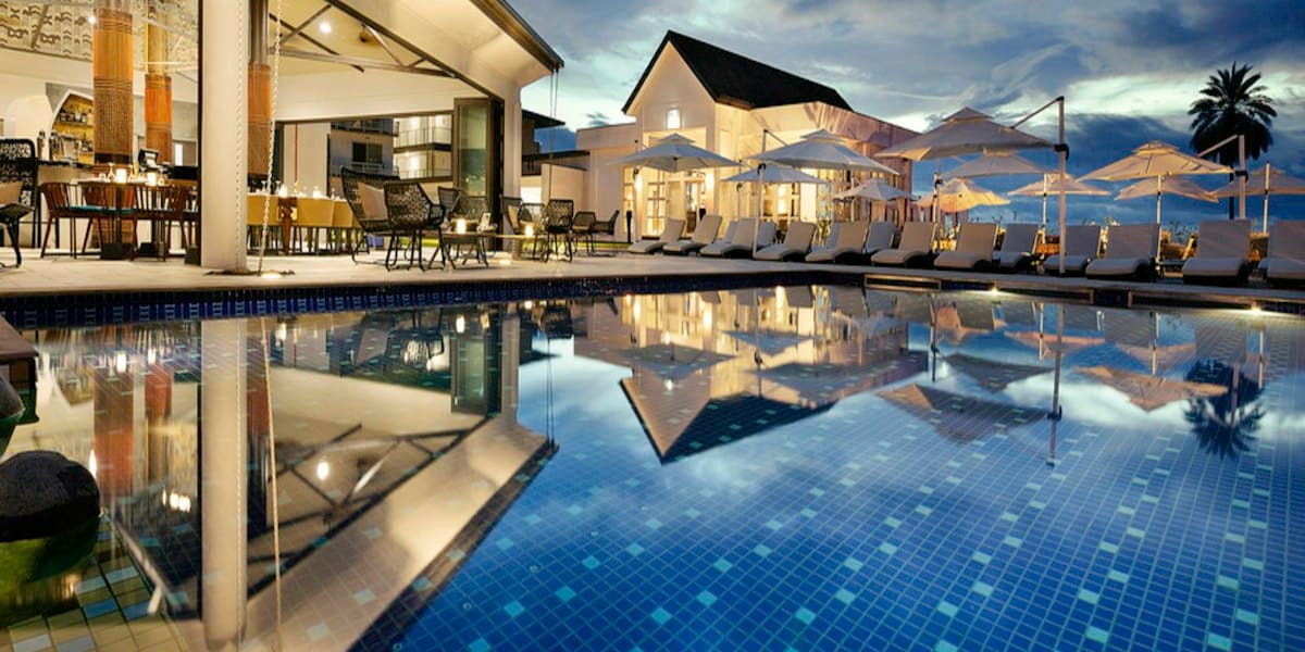 A lit resort pool deck with lounge chairs, umbrellas and a palm tree, reflected in the calm tiled pool water at dusk.