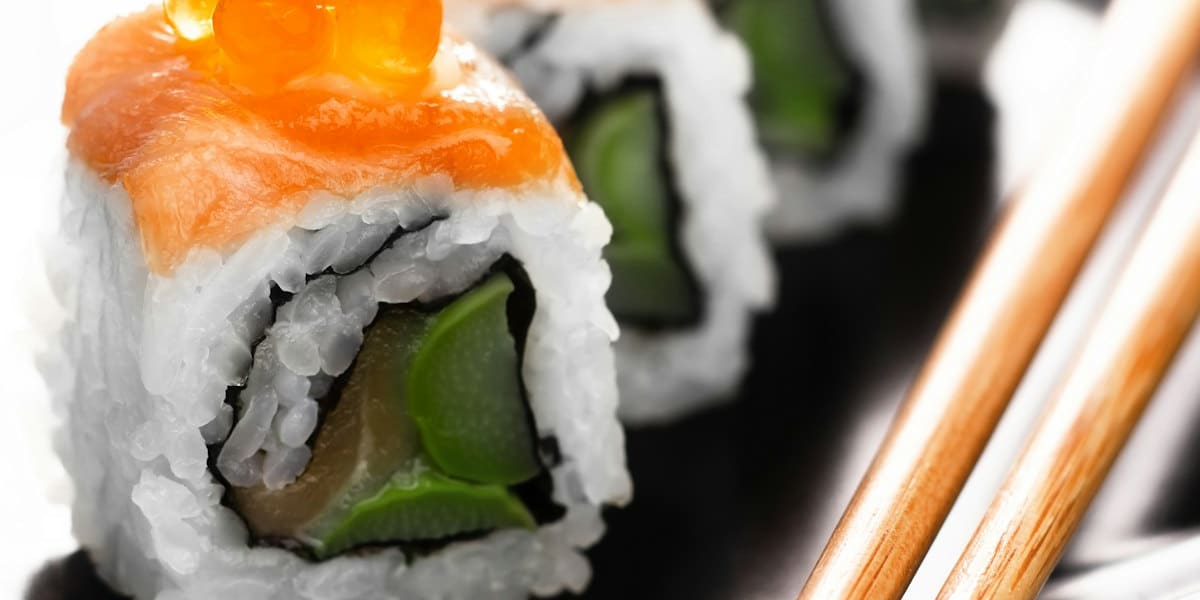 Row of sushi rolls topped with salmon and orange roe on a black plate next to wooden chopsticks
