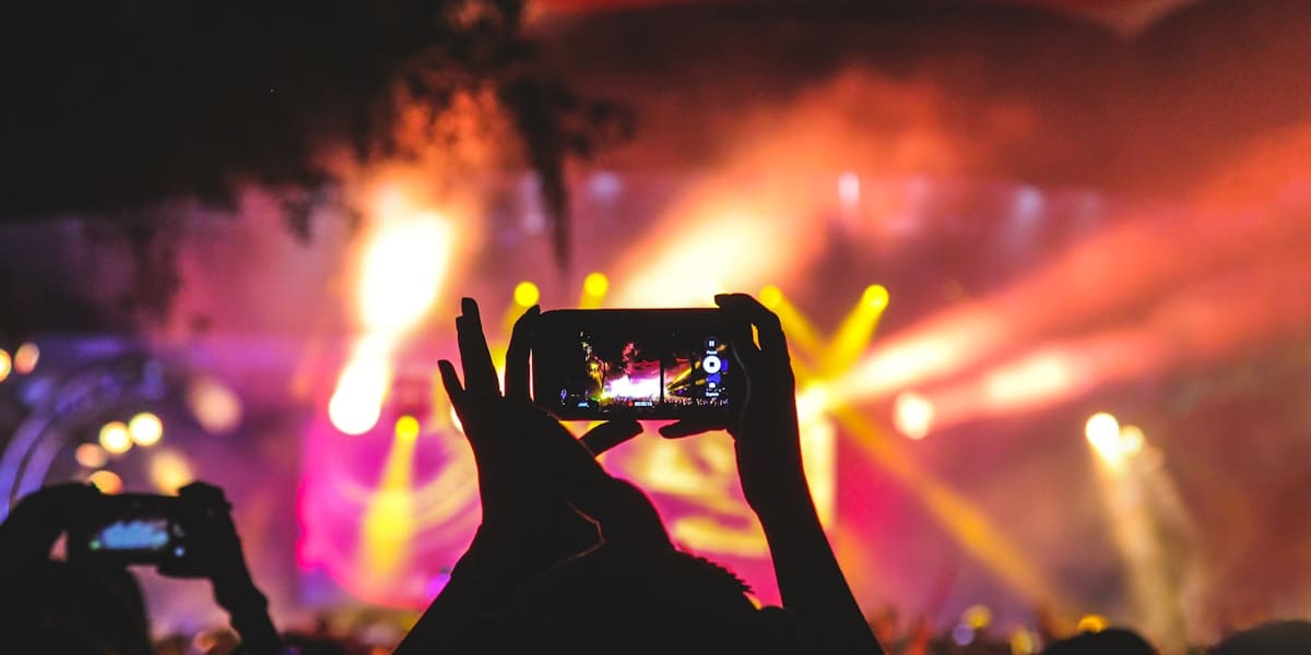 Silhouetted concertgoers raising phones to film a brightly lit stage with orange and pink spotlights at night.