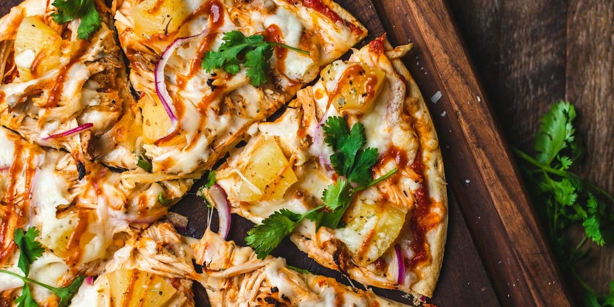 Sliced BBQ chicken and pineapple flatbread pizza with red onion and cilantro on a wood board