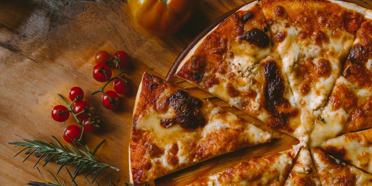 A cheese pizza sliced into wedges on a wooden board, with one slice pulled out, beside cherry tomatoes and a rosemary sprig.