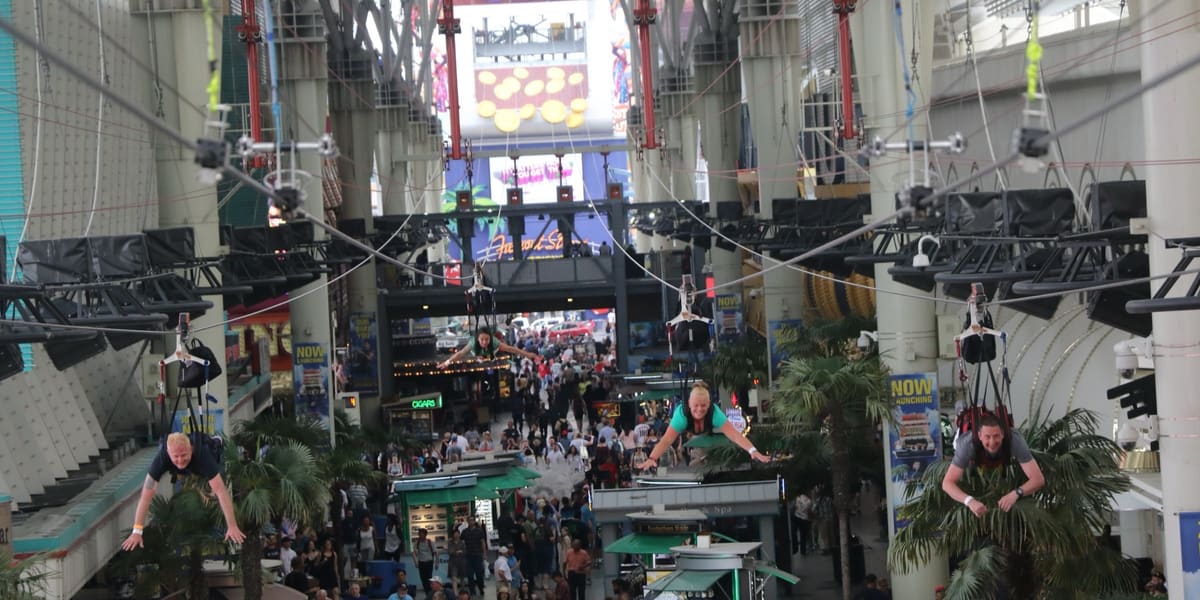 Zip line riders flying head-first above a crowded covered pedestrian mall beneath the SlotZilla tower