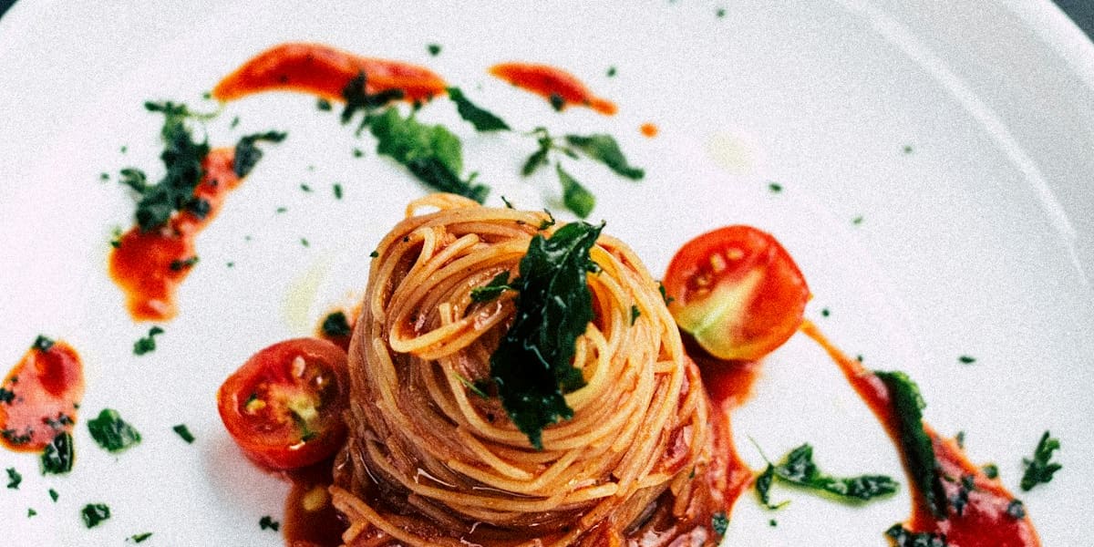 A twisted nest of spaghetti in tomato sauce plated on a white dish, garnished with basil and halved cherry tomatoes.