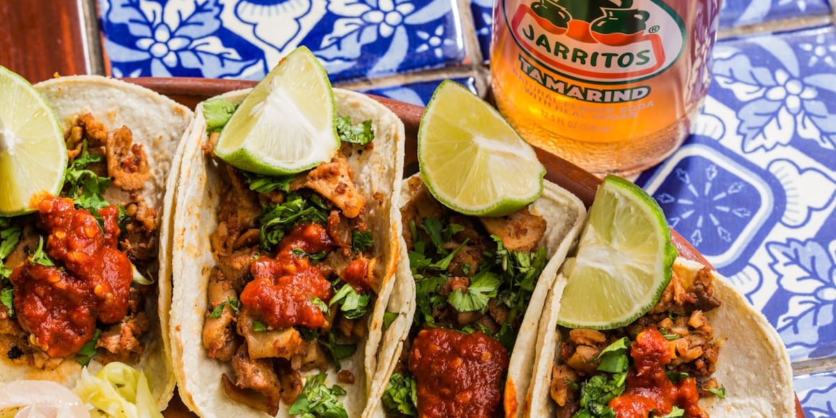 Four meat street tacos topped with salsa, onion and cilantro, served with lime wedges beside a bottled tamarind soda on patterned tile.